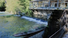 Historic wooden waterwheel and stone boat planter filled with flowers on the emerald green Sorgue River in Fontaine-de-Vaucluse, Provence.