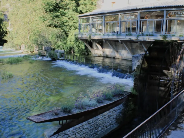 Historic wooden waterwheel and stone boat planter filled with flowers on the emerald green Sorgue River in Fontaine-de-Vaucluse, Provence.