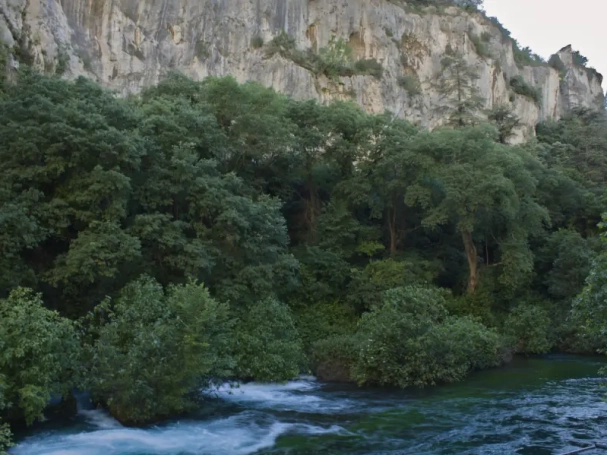 Long exposure landscape photography of the massive limestone cliffs and dense woodland canyon overlooking the emerald waters of the Sorgue river in Fontaine-de-Vaucluse, France.